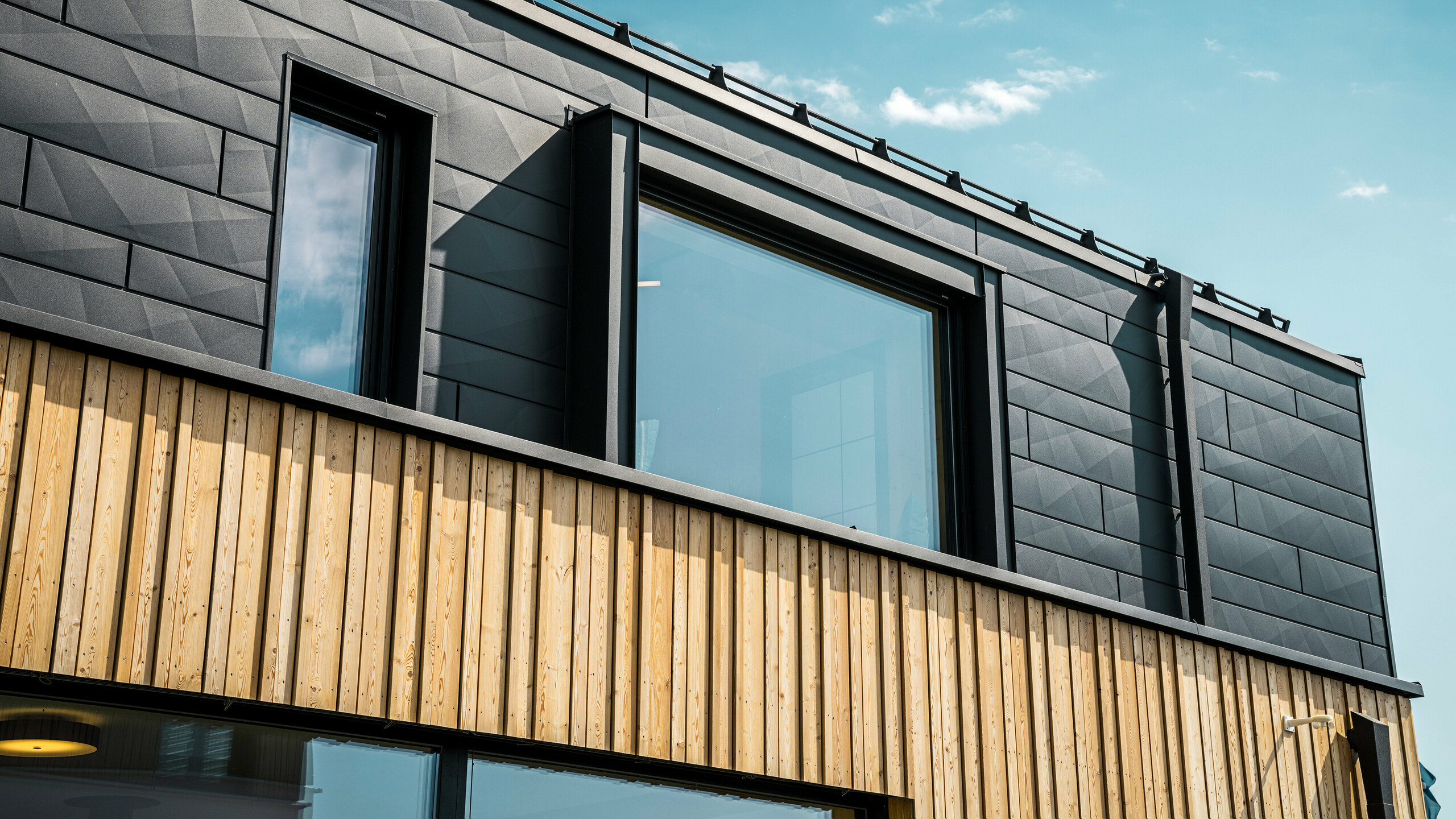 Detailný pohľad na horné poschodie vzorového domu Autark v centre prefabrikovaných domov Blaue Lagune, Vösendorf (AT). Fasáda je tvorená hliníkovými panelmi PREFA Siding.X v čiernej farbe P.10, ktoré zaujímavým spôsobom vynikajú svojím špeciálnym vzhľadom s rôznymi ohybmi. Pod nimi sa nachádza štýlové drevené obloženie, ktoré vytvára harmonický kontrast s tmavým hliníkom. Veľké okná prepúšťajú do interiéru veľa svetla a zdôrazňujú moderný dizajn.