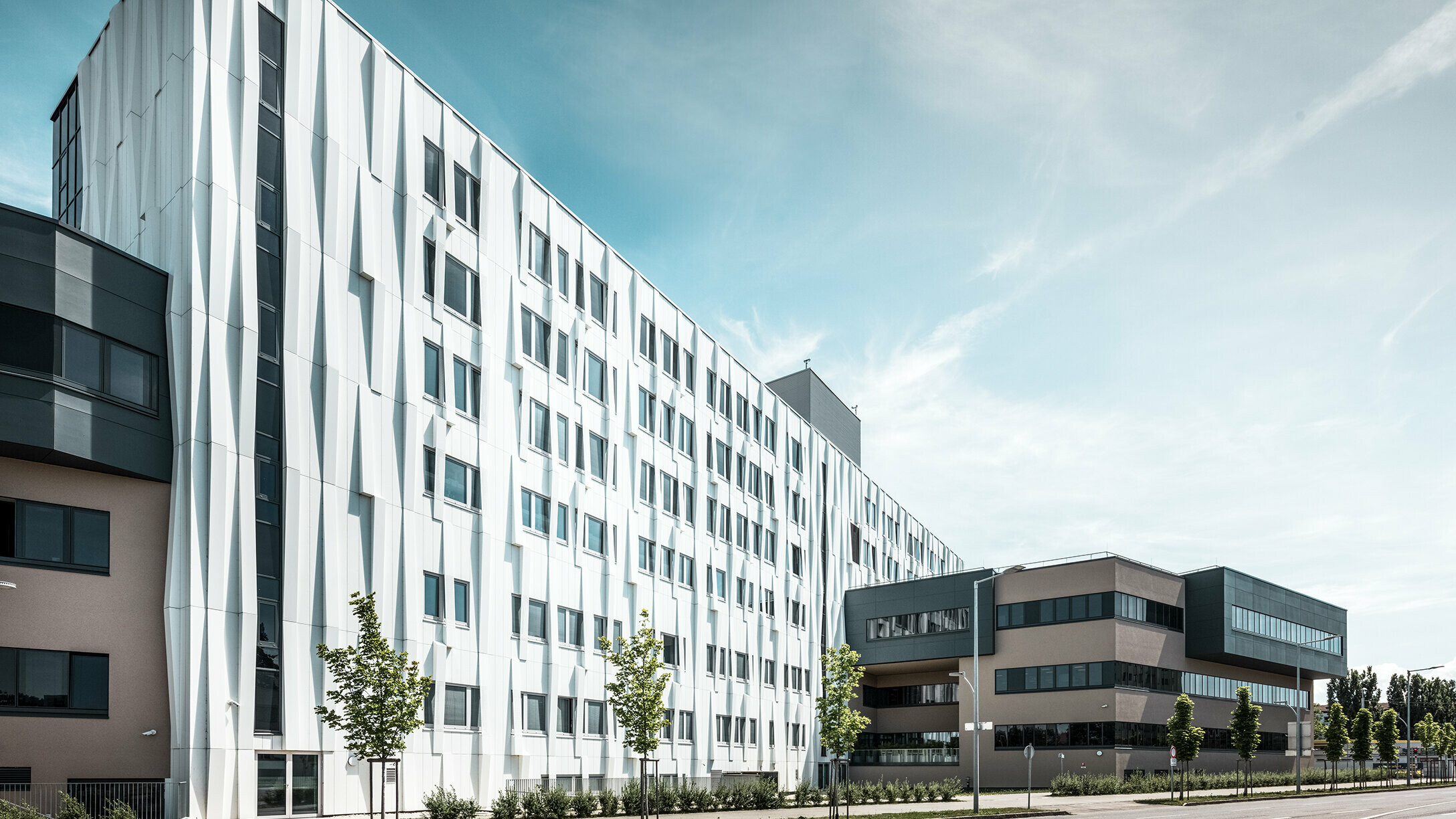 Bočný pohľad na univerzitnú kliniku v St. Pölten, Dolné Rakúsko. Robustná hliníková fasáda robí z budovy pútavý objekt v meste. V popredí je vidieť ulicu s niekoľkými stromami.