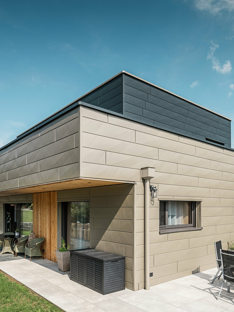 Moderný rodinný dom v rakúskom Wippenhame s dvojfarebnou hliníkovou fasádou PREFA pozostávajúcou z extravagantných fasádnych panelov Siding.X v bronzovej a antracitovej farbe. Fasáda sa vyznačuje jasne rozpoznateľnými, elegantnými líniami a ohyby. Harmonicky integrovaný strešný odvodňovací systém PREFA so zberným boxom na vodu a odvodňovacím potrubím je jasne viditeľný a dokonale zapadá do celkového architektonického konceptu. V popredí je vidieť útulnú terasu so záhradným nábytkom, ktorá podčiarkuje moderný dizajn novostavby.