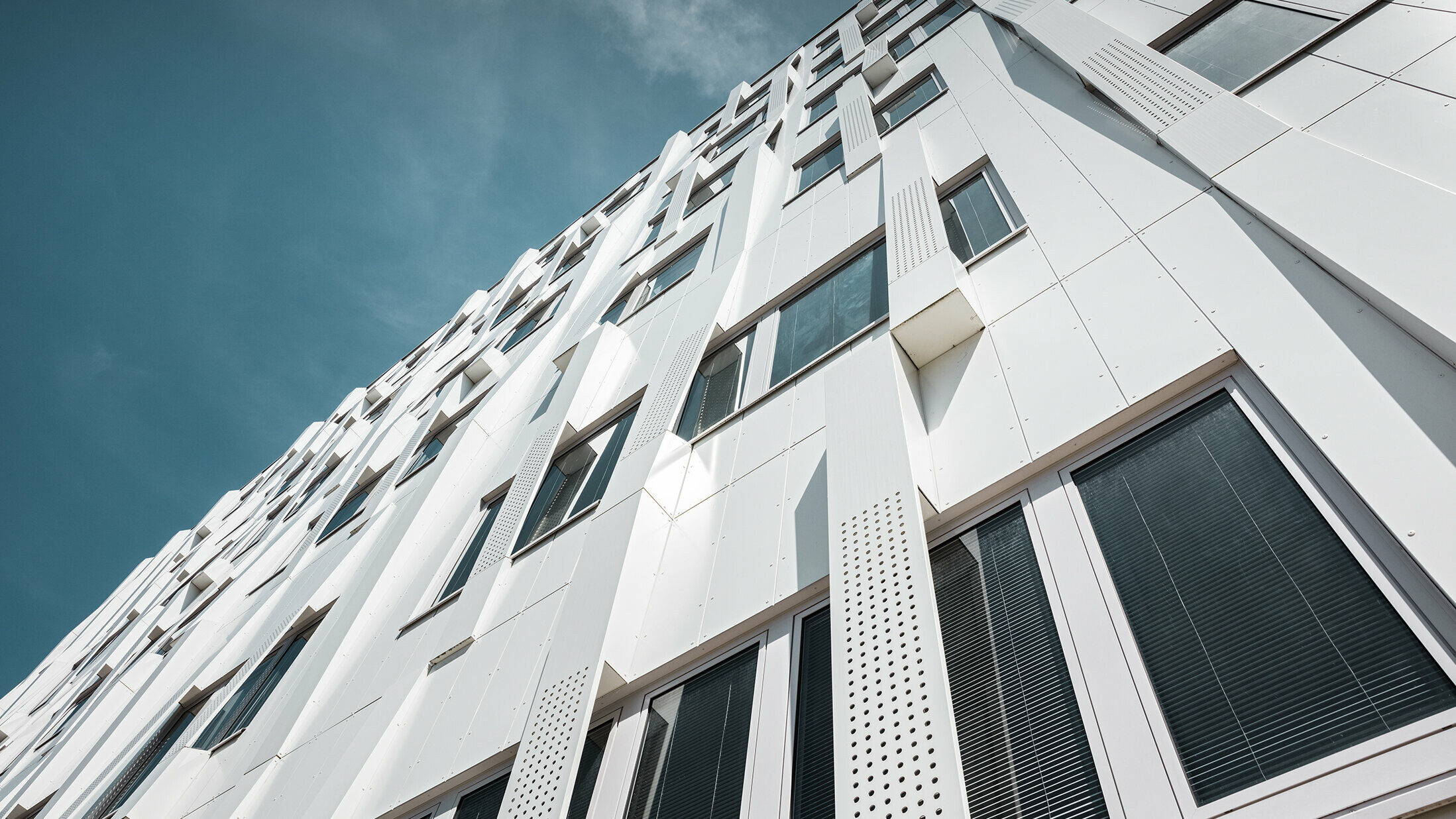 Nahaufnahme der 3D-Fassade mit gelochten Aluminium Verbundplatten-Kassetten und normalen PREFA Verbundplatten in Reinweiß. Die Kassettenfassade von PREFA macht das Gebäude zu einem Blickfang.