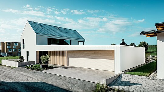 Na obrázku je moderný rodinný dom vo Freistadte, ktorý sa vyznačuje jednoduchými geometrickými tvarmi a kombináciou čisto bielej hliníkovej fasády PREFALZ a drevených obkladov. Drevený obklad na garážových dverách vytvára teplý, prirodzený kontrast a dokonale zapadá do moderného dizajnu. Strecha, tiež pokrytá PREFALZ, zaisťuje hladký povrch odolný voči poveternostným vplyvom. Táto kombinácia ľahko udržiavateľného hliníka a dreva vytvára esteticky príťažlivé a funkčné riešenie pre udržateľnú architektúru. Rozľahlý zelený exteriér a jasná obloha v pozadí podčiarkujú modernú kvalitu života, ktorú tento dom ponúka.