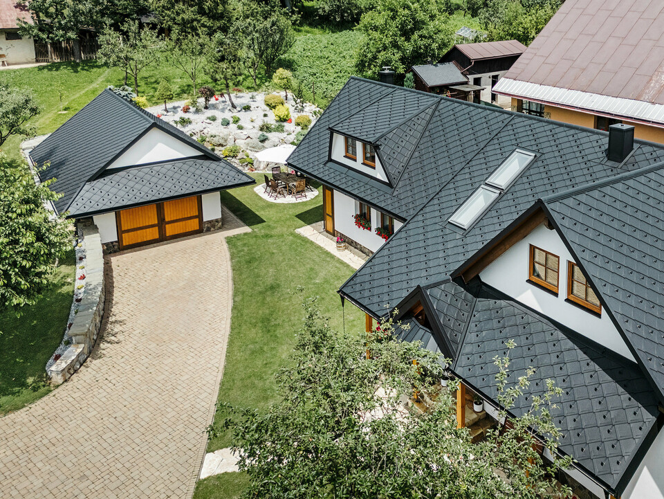 Letecký pohľad na tradičný rodinný dom v Starej Bystrici na Slovensku, pokrytý strešnými šablónami PREFA 29 × 29 vo farbe P.10 čierna. Strecha zaujme elegantným pultovým vzhľadom a harmonickým začlenením vikierov a strešných okien. Okolitá zeleň, udržiavaná záhrada a vydláždená príjazdová cesta podčiarkujú príťažlivý celkový vzhľad budovy.