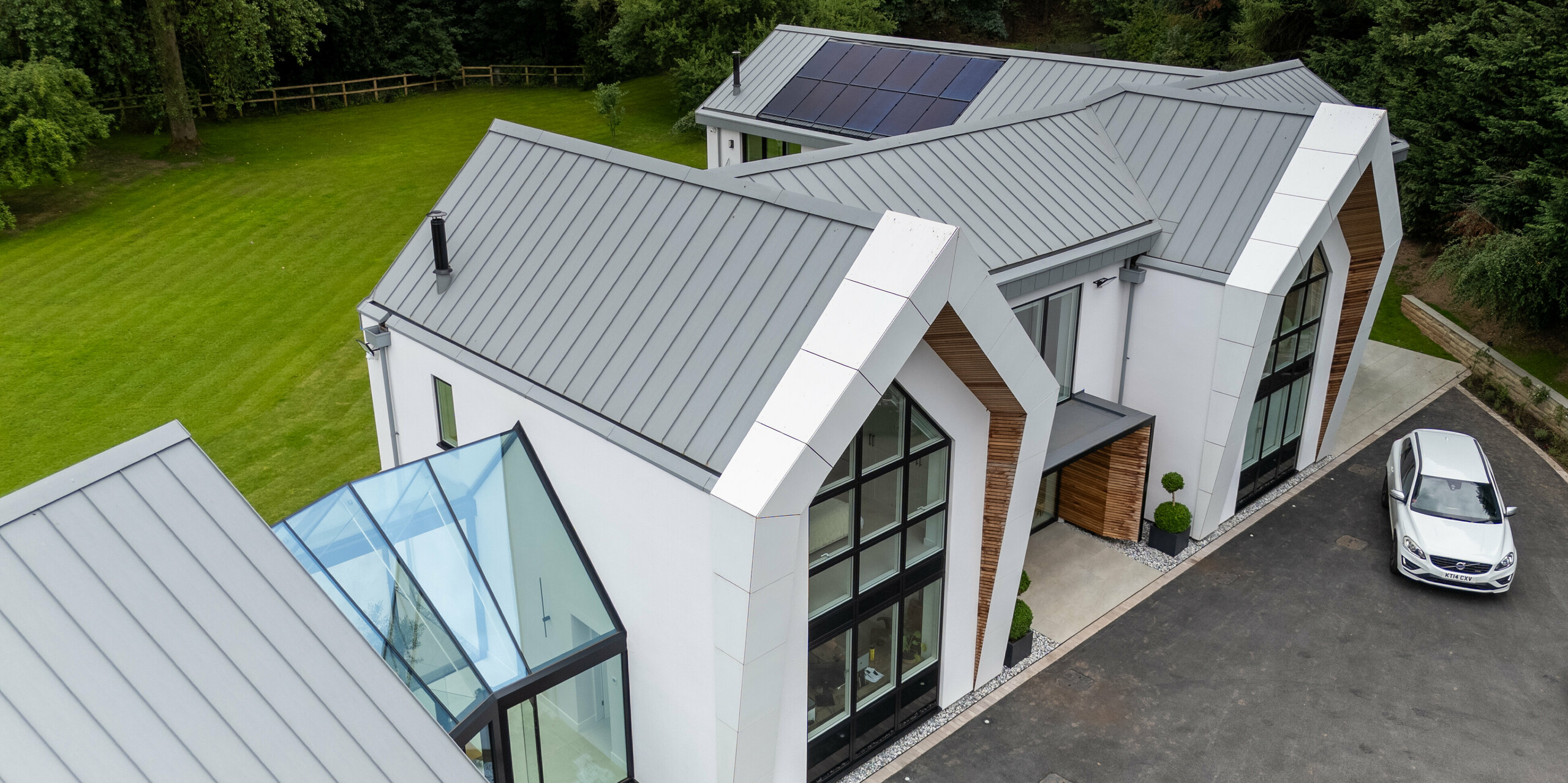 Das Bild zeigt die moderne Villa in Draycott in the Clay aus einer eindrucksvollen Perspektive. Ihr elegantes PREFALZ-Dach in Patinagrau ist ein echter Blickfang. Die klaren Stehfalzstrukturen verleihen der Architektur ein markantes, modernes Erscheinungsbild und fügen sich harmonisch in die grüne Umgebung ein. Das Bild hebt zudem die interessante Kombination von Glasflächen und Holzverkleidung an der Fassade hervor, die zusammen mit dem langlebigen PREFALZ-Dach für ästhetische Raffinesse sorgt. Die Photovoltaikanlage auf dem Dach unterstreicht die nachhaltige Ausrichtung des Hauses.