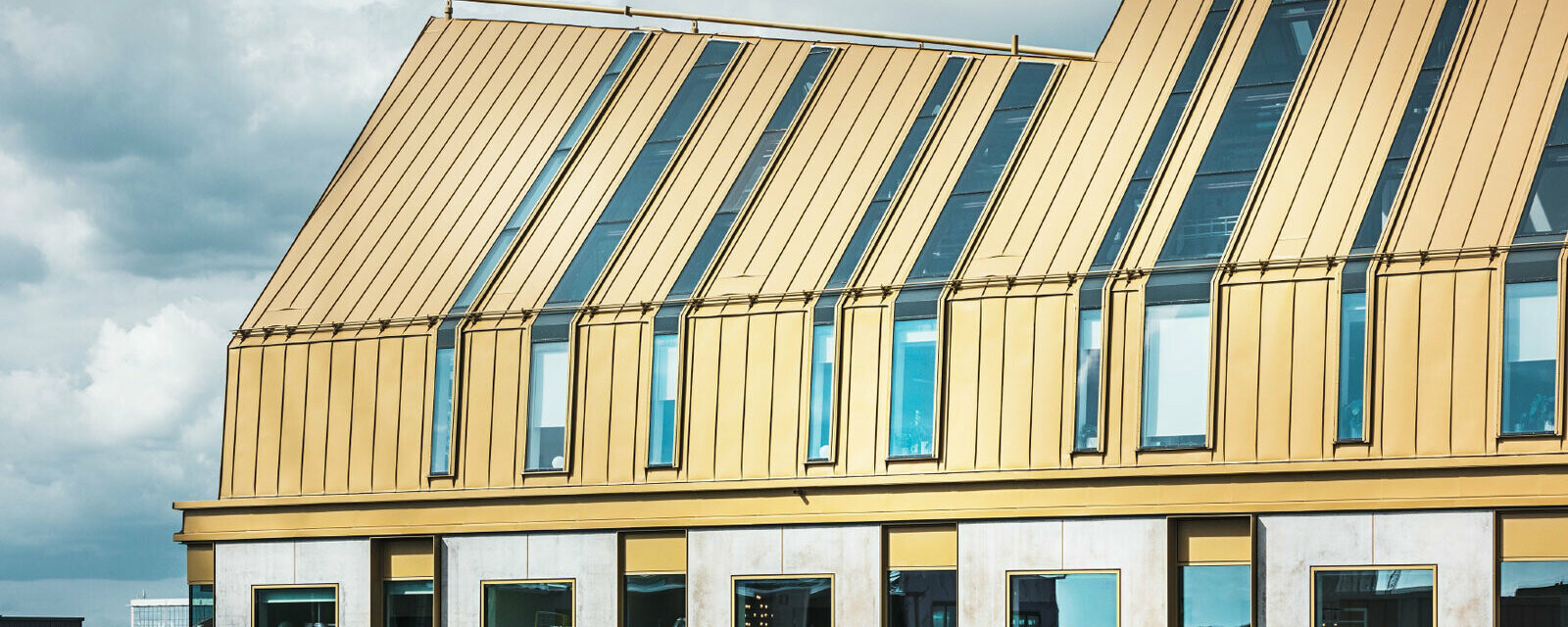 Moderner, mehrgeschossiger Bürobau mit charakteristischer goldfarbener Stehfalz‑Metalldach‑ und Fassadenverkleidung, vertikal gegliederte Fensterflächen und großflächige Dachflächenfenster, klare Linienführung und zeitgenössische Materialität vor bewölktem Himmel – repräsentativ für ein funktionales, anspruchsvolles Bürogebäude mit präziser Aluminiumverarbeitung und starker architektonischer Präsenz.