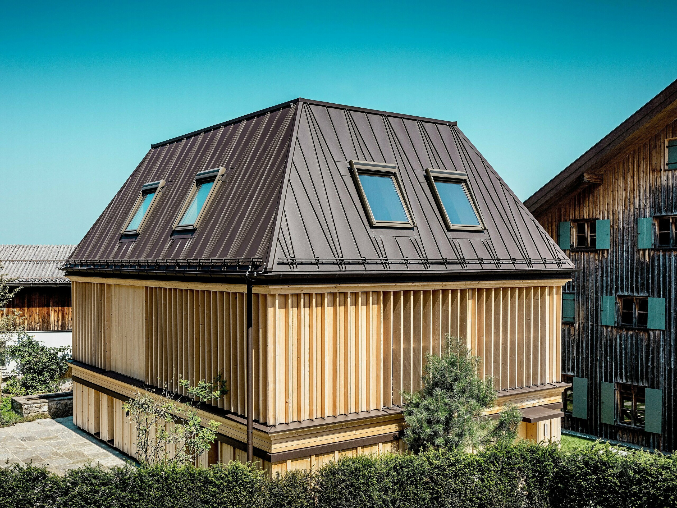 Novo postavený „kúpeľný dom“ hotela Hirschen v Schwarzenbergu za denného svetla. V popredí upúta pozornosť výrazná strecha PREFA so stojatými falcami v odtieni P.10 orieškovohnedá, ktorej atypicky umiestnené falce dodávajú valbovej streche dynamický, moderný vzhľad. Sklon strechy 60 stupňov podčiarkuje výraznú architektúru. Fasáda sa skladá z vertikálne usporiadaných drevených lamiel po celej výške poschodia, ktoré efektívne vytvárajú hru svetla a tieňa a zároveň poskytujú funkčnú ochranu pred slnkom. Kombinácia tradičného tvaru budovy a inovatívneho výberu materiálov vytvára zaujímavý kontrast k pamiatkovo chránenej existujúcej budove v pozadí, ktorá je so svojou zvlnenou drevenou fasádou typická pre región Bregenzerwald.