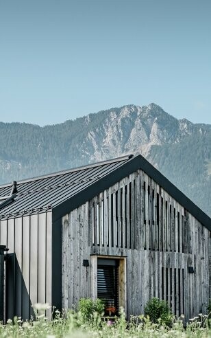 Moderner, funktionaler Wohnbau mit einer Kombination aus Holzfassade und PREFA Band- und Blechmaterial für das Dach. Die klare Architektur wird durch die elegante, metallische Dach- und Fassadenverkleidung unterstrichen, die ideal für anspruchsvolle Projekte ist. Das Bild veranschaulicht die vielseitigen Anwendungsmöglichkeiten der PREFA Produkte im Bereich Bänder und Bleche.