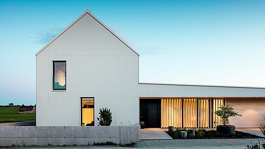 Na obrázku je impozantný rodinný dom vo Freistadte, ktorý sa vyznačuje výraznou geometrickou architektúrou. Fasáda budovy je z presne ohýbaného, čisto bieleho PREFALZ, ktorý dodáva domu modernú eleganciu. Vertikálna štruktúra fasády a čistý, minimalistický dizajn podčiarkujú vysokú kvalitu použitých materiálov a zaisťujú odolný povrch odolný voči poveternostným vplyvom. Strmá sedlová strecha, ktorá je tiež pokrytá plechom PREFALZ, chráni pred živlami a zároveň zaisťuje harmonický celkový vzhľad. Vďaka jasnému dizajnu a použitiu hliníka sú optimálne splnené estetické aj funkčné požiadavky. Svetlom zaplavené okná a otvorený vstupný portál pôsobia prívetivo a vytvárajú zaujímavý kontrast s moderným dizajnom fasády. Rozhodnutie pre PREFALZ podčiarkuje architektonické nároky a predstavuje udržateľné, ľahko udržiavateľné riešenie pre dlhotrvajúce stavebné projekty.