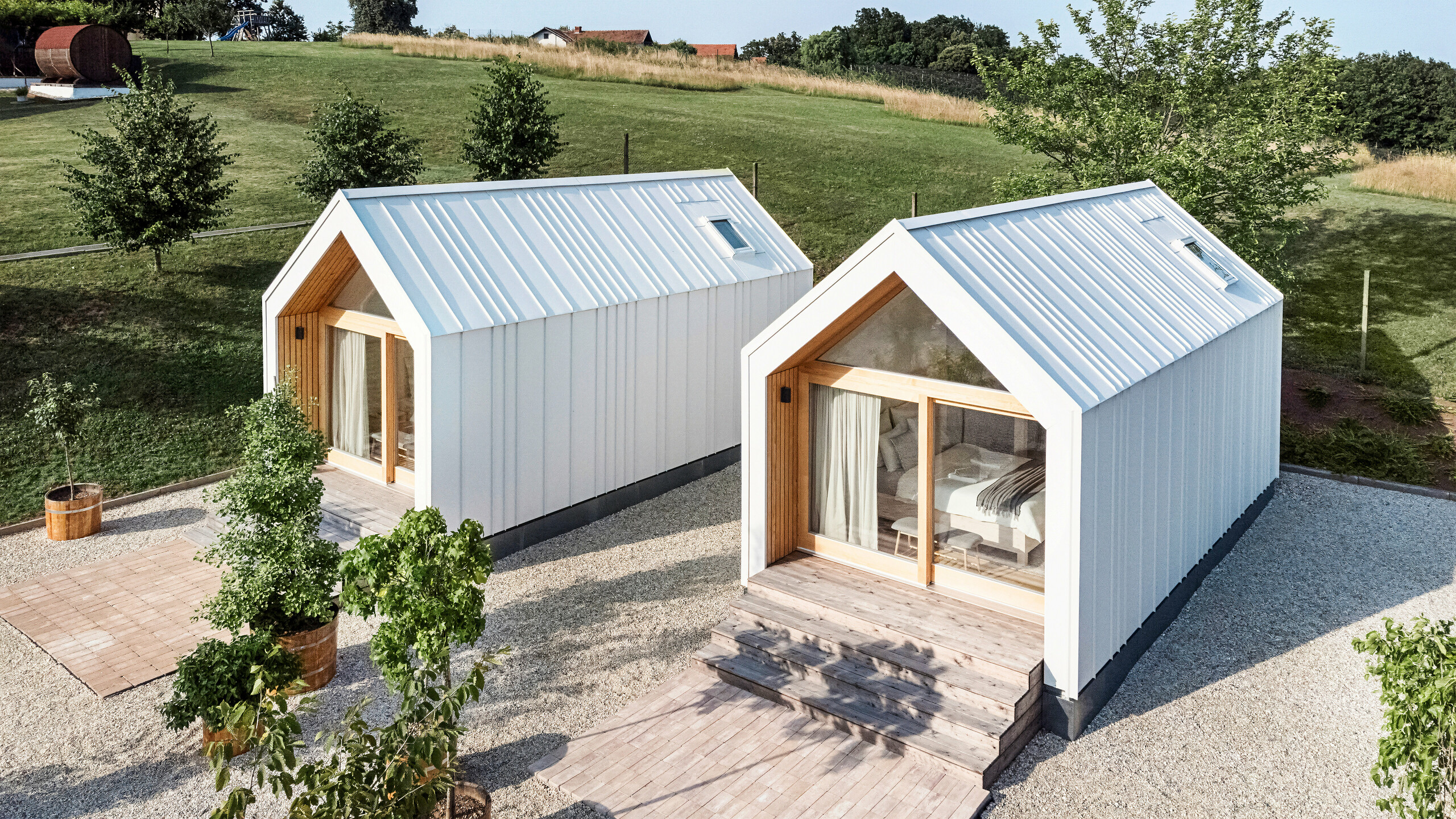 Pohľad na priečelie dvoch malých domčekov v rekreačnom komplexe Pri Momi v Kančevci, Slovinsko. Mikrodomčeky boli postavené s použitím drevenej rámovej konštrukcie a dokončené robustnou strechou PREFALZ a fasádnym obkladom vo farbe P.10 čistá biela. Moderné, minimalistické budovy kombinujú odolný a zároveň estetický hliníkový plášť s veľkými sklenenými priečeliami a prírodným drevom, zasadené do zelenej krajiny. Ku vchodom vedú veľkorysé drevené schody, ktoré vytvárajú útulnú a príjemnú atmosféru.