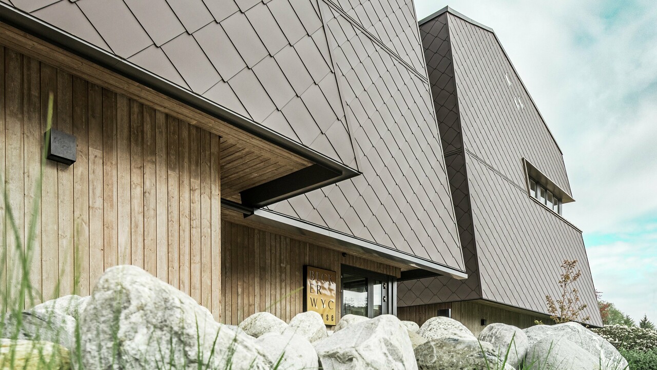 Bočný pohľad na hotel, fasáda prízemia je vertikálne obložená drevom tmavej farby. Nad ním je fasádny výstupok, odtiaľ je fasáda obložená hliníkovými panelmi v orieškovohnedej farbe.