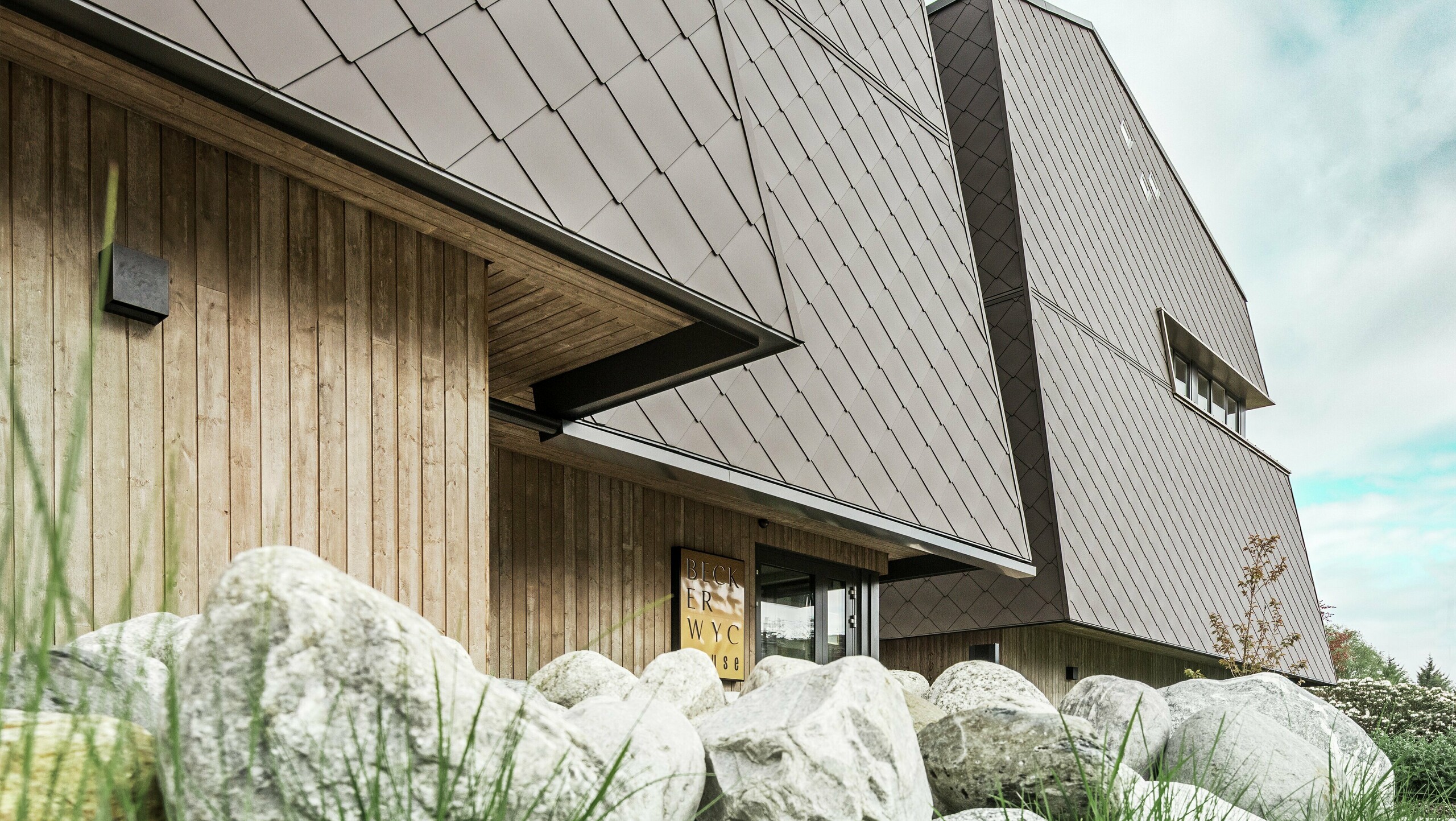 Bočný pohľad na hotel, fasáda prízemia je vertikálne obložená drevom tmavej farby. Nad ním je fasádny výstupok, odtiaľ je fasáda obložená hliníkovými panelmi v orieškovohnedej farbe.