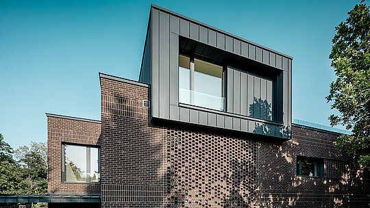 Obrázok zachytáva výrazné riešenie fasády modernej viacgeneračnej vily v Budapešti, kde hliník PREFALZ v odtieni P.10 antracit vytvára jasný architektonický akcent. Vystupujúci kubus s precíznymi stojatými falcami vytvára silný kontrast k štruktúrovanej tehlovej fasáde pod ním. Vďaka bočnému slnečnému svetlu vzniká dynamická hra tieňov a línií, ktorá zvýrazňuje kvalitné spracovanie a dlhú životnosť hliníkového riešenia PREFA. Kombinácia kovu a klinkeru podčiarkuje moderný, kontrastný charakter budovy.