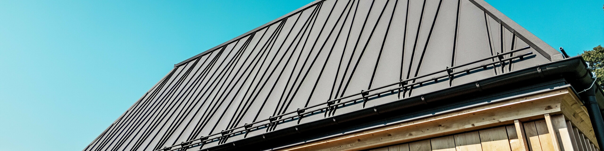 Detailný pohľad na výnimočnú strechu PREFA so stojatými falcami na „kúpeľnom dome“ pri hoteli Hirschen v Schwarzenbergu. Strecha vo farbe P.10 orieškovohnedá zaujme svojimi výraznými, čiastočne diagonálnymi stojatými falcami, ktoré budove dodávajú dynamický, moderný vzhľad. Čisté línie a sklon strechy 60 stupňov vytvárajú silný geometrický efekt. Pod strechou je viditeľná fasáda s vertikálne usporiadanými drevenými lamelami, ktoré nanovo interpretujú tradičný spôsob výstavby. Bezoblačná obloha a priame slnečné svetlo zdôrazňujú vysokú kvalitu povrchu materiálu PREFALZ a precíznu prácu klampiara. Obrázok pôsobivo zachytáva spojenie inovácie, remeselného umenia a regionálneho štýlu.