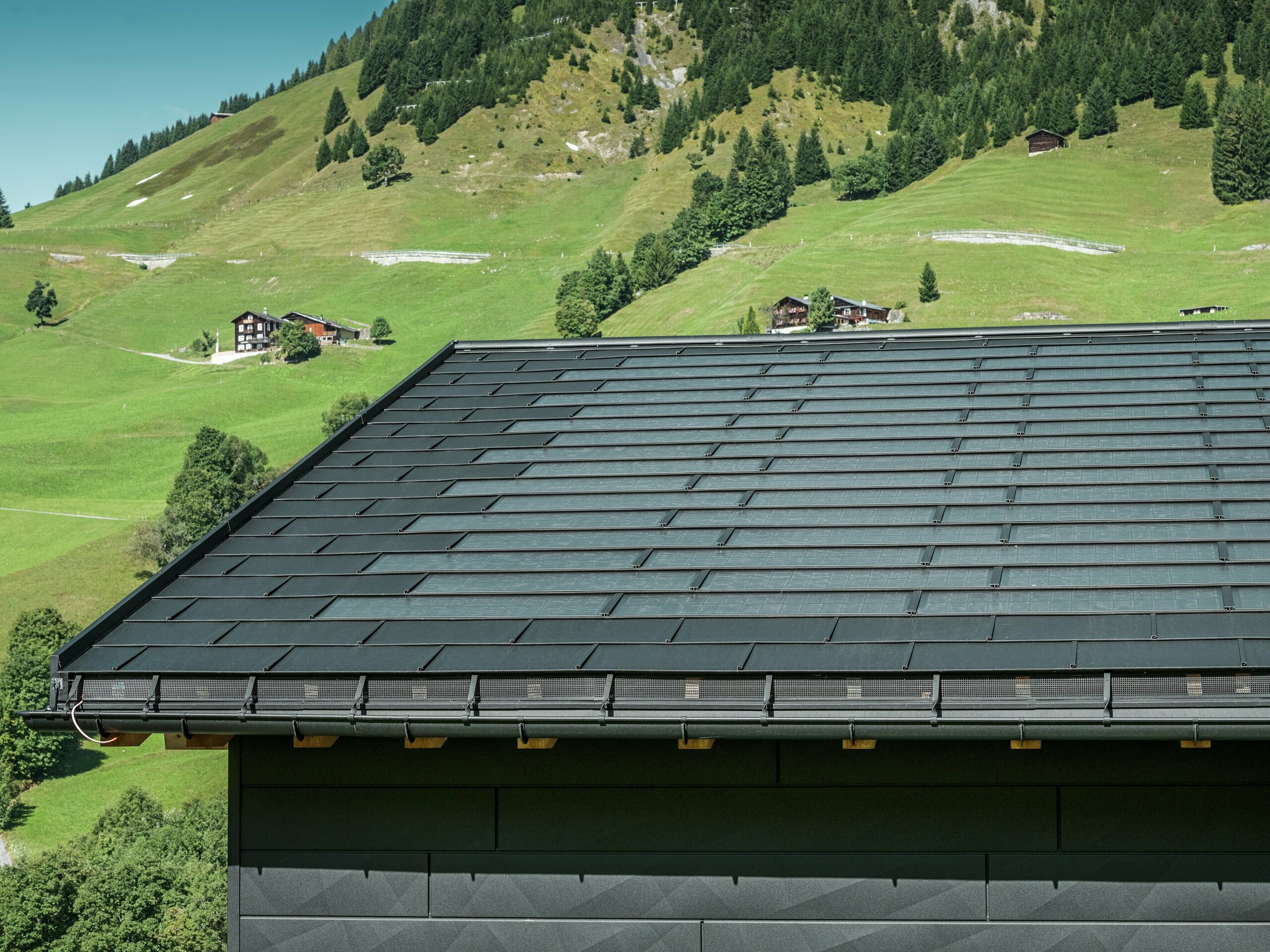 Detailný pohľad na odkvap moderného rodinného domu v alpskom prostredí Ascharina vo Švajčiarsku. Strecha bola pokrytá integrovanými solárnymi panelmi PREFA a tradičnými strešnými panelmi R.16, obe v čiernej farbe P.10. Spoľahlivé odvodnenie strechy zabezpečujú odvodňovacie žľaby a zvody PREFA. V spodnej časti obrázku sú viditeľné nezameniteľné fasádne panely PREFA Siding.X. Čisté línie hliníkových produktov podčiarkujú moderný dizajn uprostred malebnej horskej krajiny.