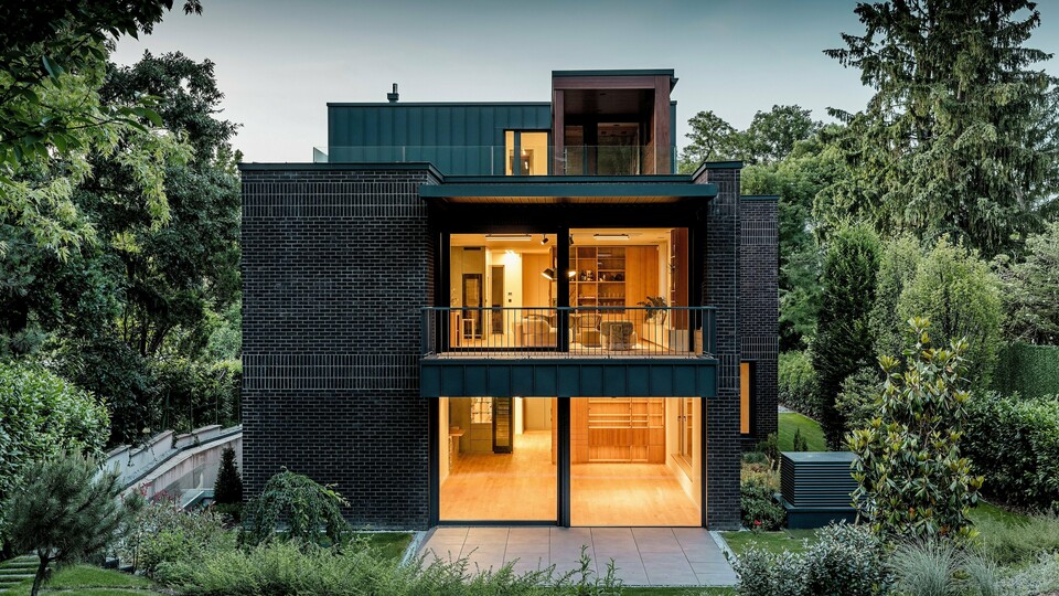 Fotografia zachytáva modernú zadnú stranu budapeštianskej viacgeneračnej vily za súmraku. Veľkorysé presklené fasády ponúkajú výhľad na príjemne osvetlené interiéry, zatiaľ čo kombinácia tmavočervenej tehlovej fasády a hliníka PREFALZ v odtieni P.10 antracit zdôrazňuje čisté architektonické línie. Presne vyhotovené uhlové stojaté falce podčiarkujú vysokú kvalitu spracovania a dlhú životnosť hliníkových produktov PREFA. Obklopená bujnou zeleňou sa budova harmonicky začleňuje do záhradnej krajiny a vyžaruje elegantný, moderný charakter.