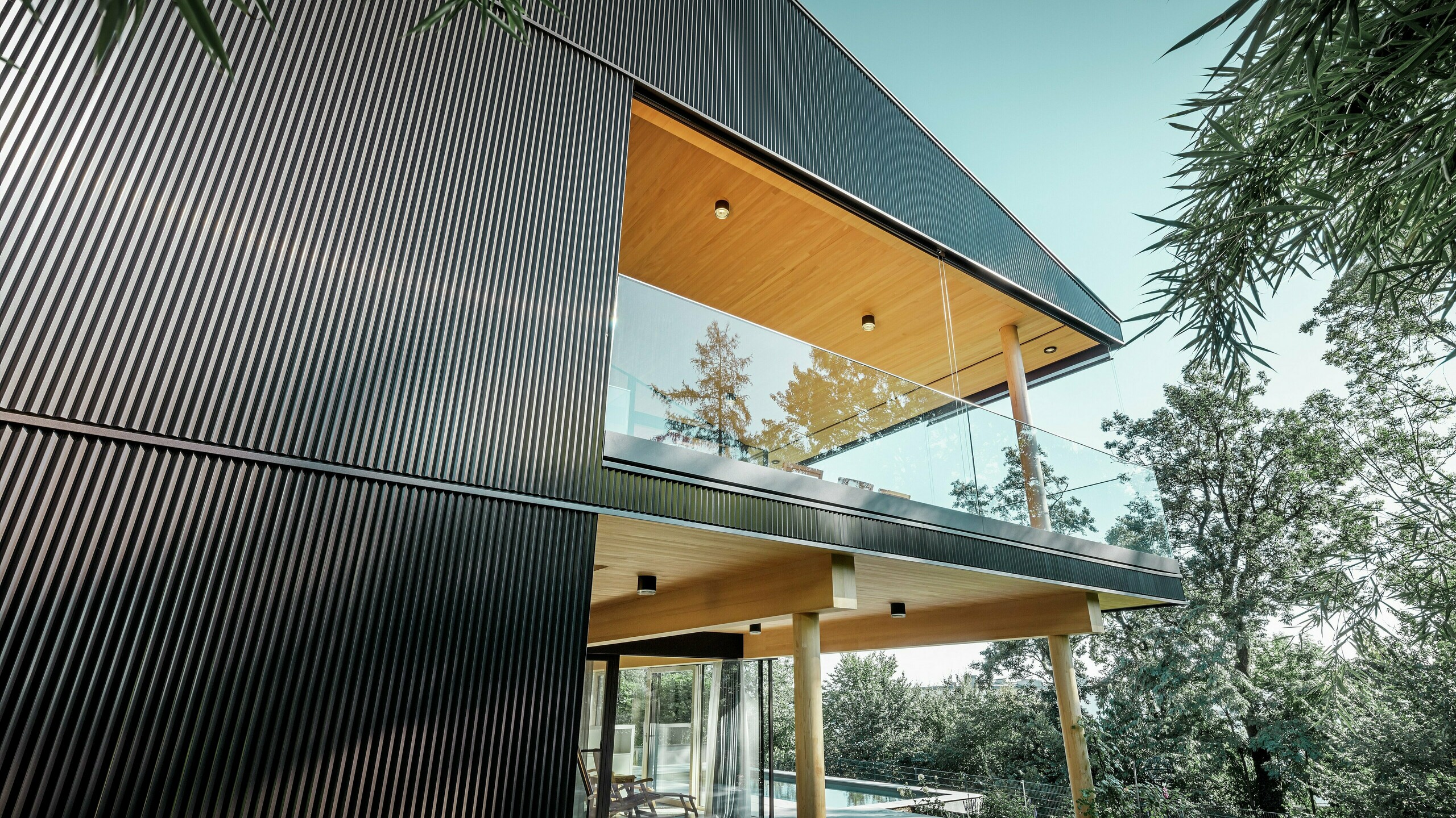 Bočný pohľad na rodinný dom, fasáda je obložená tmavým prelamovaným profilom PREFA. Vidno terasu a priestranný balkón. Strop alebo podhľad je riešený drevenými panelmi.