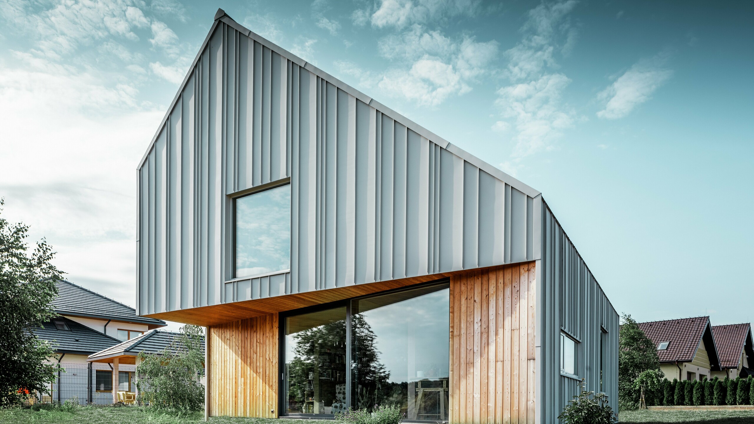 Moderný rodinný dom so šikmou strechou bez odkvapov a hliníkovou fasádou od spoločnosti PREFA v bielej farbe. Drevená fasáda je kombinovaná so zvislými drevenými panelmi na prízemí. 