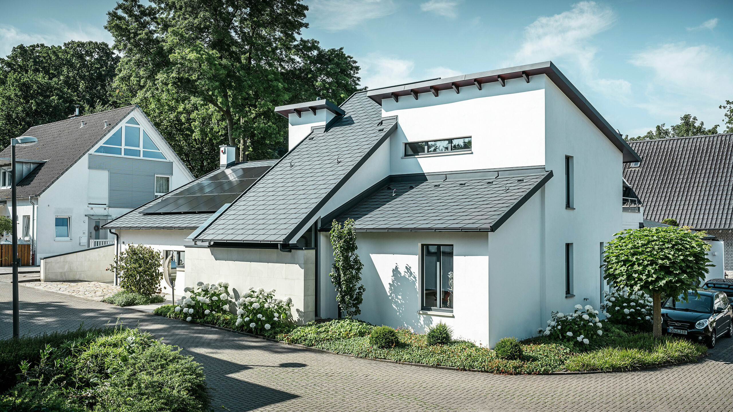 Moderný rodinný dom s bielou omietkovou fasádou a tmavou šikmou strechou PREFA z hliníka. Na streche sú integrované fotovoltaické moduly a hliníkové odvodňovacie systémy v antracitovej farbe. Budova sa nachádza v udržiavanej obytnej štvrti so zelenými predzáhradkami, dláždenou príjazdovou cestou a okolitými rodinnými domami.
