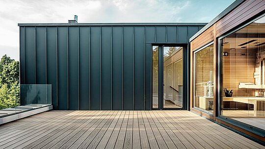 Záber zachytáva rozsiahlu strešnú terasu budapeštianskej viacgeneračnej vily, ktorej fasáda je obložená hliníkom PREFALZ v odtieni P.10 antracit. Precízne zvisle vedené stojaté falce vytvárajú čisté, moderné línie, ktoré dokonale ladia s teplým drevom susednej sauny. Prirodzené slnečné svetlo zvýrazňuje elegantný kontrast materiálov a podčiarkuje vysokú kvalitu a dlhú životnosť riešenia PREFA. Priestranná terasa otvára výhľad do zelene a zdôrazňuje moderný, komfortne orientovaný charakter budovy.