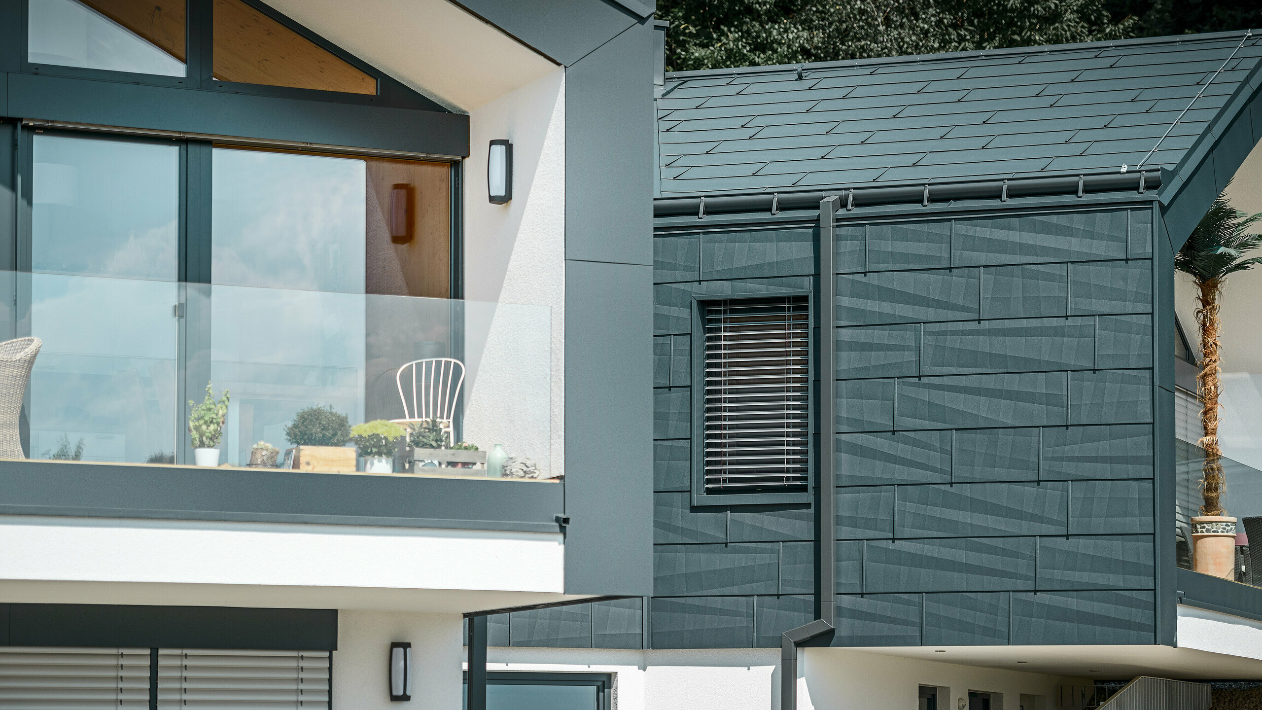 Na obrázku je moderný dvojdom so štýlovou fasádou z hliníka. Fasáda je v tmavom antracitovom odtieni a má štruktúrovaný povrch vďaka strešným a fasádnym panelom PREFA FX.12. Na streche bola namontovaná nástrešný žľab a štvorcový zvod ako dažďový zvod od spoločnosti PREFA. Na hornom poschodí je balkón so skleneným zábradlím, zatiaľ čo na spodnom poschodí sú okná s lamelovými žalúziami. Architektúra pôsobí jasne a minimalisticky a v pozadí sú vidieť stromy, ktoré vytvárajú prírodnú atmosféru.