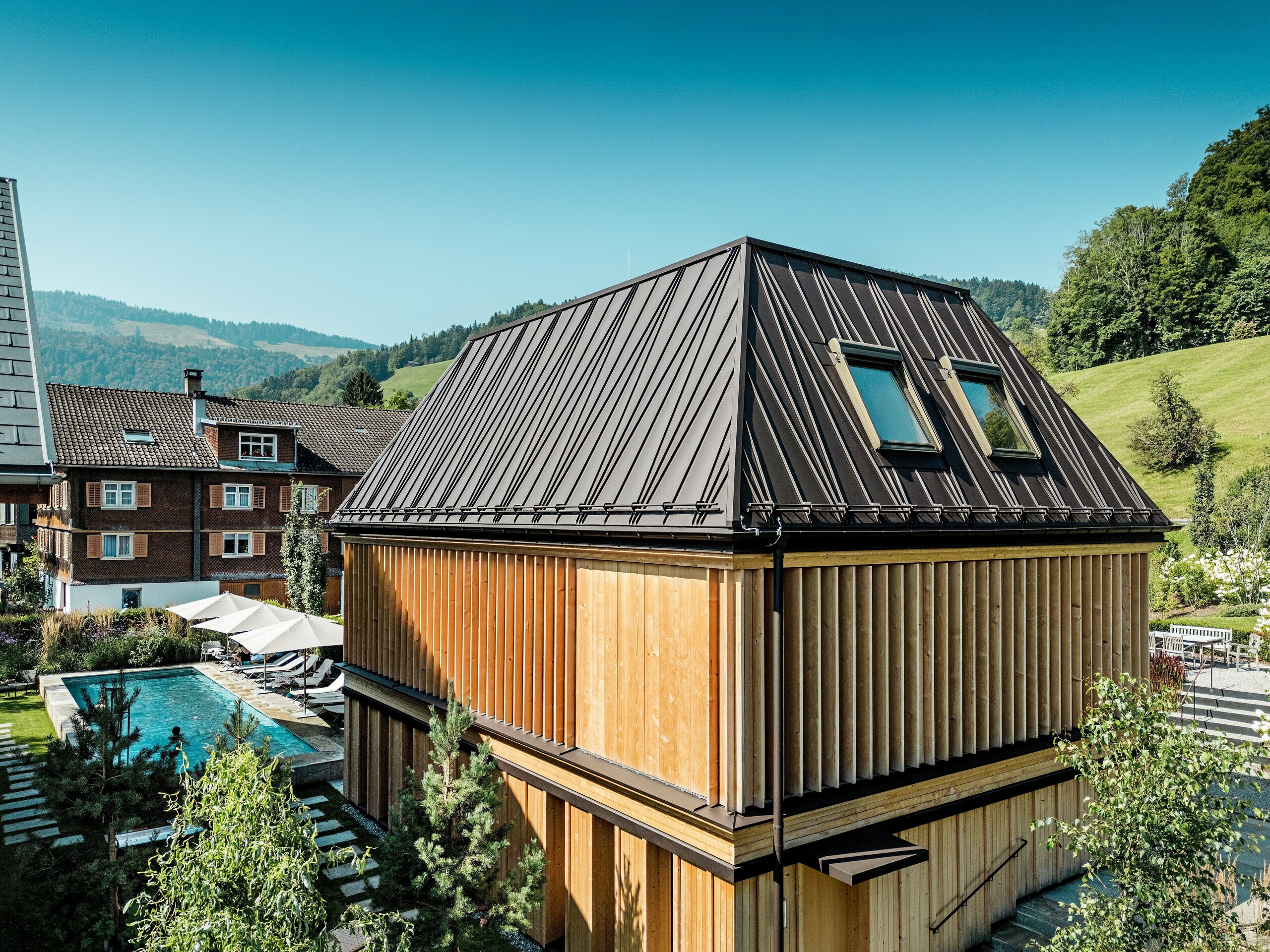 Moderný „kúpeľný dom“ hotela Hirschen v Schwarzenbergu za slnečného dňa, zasadená do zelenej kopcovitej krajiny Bregenzerwald. Budova zaujme svojou nápadnou strechou PREFA so stojatými falcami v odtieni P.10 orieškovohnedá s výrazným šikmým vzorom falcov a dvoma strešnými oknami. Fasáda sa skladá z vertikálnych drevených lamiel po celej výške poschodia, ktoré flexibilne regulujú svetlo, tieň a ochranu pred pohľadmi. Na ľavej strane obrázku je vidieť vonkajší bazén s ležadlami – miesto pokoja a oddychu. Kombinácia vysokokvalitnej architektúry, regionálnych materiálov a premysleného dizajnu harmonicky zapadá do tradičného prostredia.