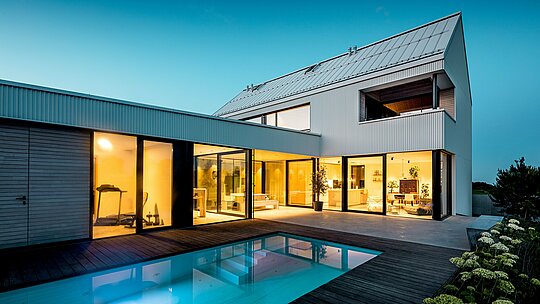 Obrázok zobrazuje moderný rodinný dom vo Freistadte s výraznou fasádou PREFALZ v čisto bielej farbe. Presne zložená hliníková fasáda dodáva budove jasnú geometrickú eleganciu, ktorá harmonicky ladí s veľkými okennými plochami a plochou strechou. Použitie PREFALZ zaručuje nielen estetickú stálosť, ale aj vysokú odolnosť voči poveternostným vplyvom a dlhú životnosť. Otvorený interiér s výhľadom na bazén dokonale dopĺňajú čisté línie a minimalistický dizajn, vďaka čomu je dom moderným a udržateľným riešením bývania.