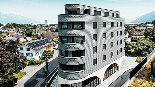 Letecký pohľad na Bernerhaus v Buchsi s výraznou trojuholníkovou kubatúrou a jemne zaoblenými balkónmi. Fasáda z PREFA šindľov v odtieni P.10 svetlošedá obklopuje budovu ako šupinatá hliníková koža a vytvára živú hru svetla a tieňa. Nad masívnym betónovým podstavcom s elegantnými arkádami pôsobí ľahká, odolná proti poveternostným vplyvom PREFA hliníková fasáda mimoriadne precízne, trvalo a s nízkymi nárokmi na údržbu – moderný pohľad upútavajúci uprostred alpského mestského prostredia.