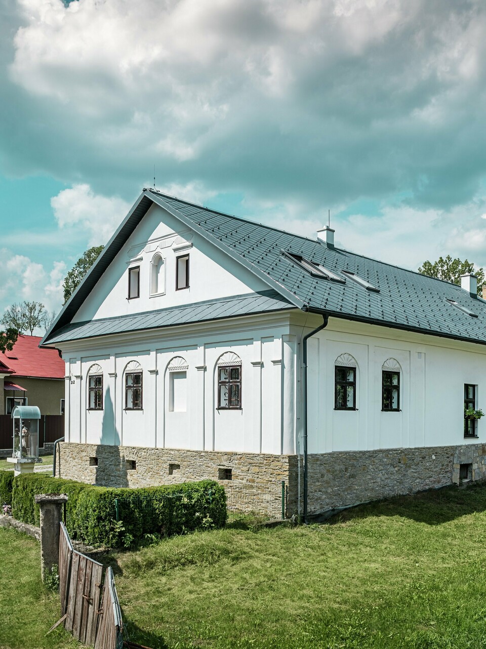 Das Bild von der Straße zeigt die Front und Seite des sanierten Handwerkerhauses in Klin, Slowakei, mit dem modernen Dach aus PREFA Dachrauten 44 × 44 in P.10 Steingrau. Der Blick auf die Gebäudeseite zeigt das elegante Satteldach und die perfekt integrierten Dachfenster, die für ein helles, offenes Ambiente sorgen. Die Dachrauten aus Aluminium ergänzen die traditionelle weiße Fassade und das Natursteinmauerwerk, was dem Haus einen zeitgemäßen und gleichzeitig klassischen Charakter verleiht. Mit ihren Vorteilen wie Langlebigkeit, Leichtigkeit und Korrosionsbeständigkeit ist die Dachlösung nicht nur ästhetisch ansprechend, sondern auch besonders funktional und nachhaltig.