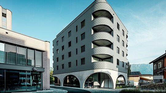 Tento moderný bernský dom v Buchsi zaujme svojím výrazne zaobleným trojuholníkovým tvarom a súvislou fasádou z PREFA šindľov v farbe P.10 svetlošedá. Šupinatý povrch hliníka zdôrazňuje mäkké hrany a hlboké zaoblenia balkónov, zatiaľ čo masívny betónový podstavec s elegantnými arkádami vytvára zaujímavý kontrast. Na pozadí Álp uprostred malej nákupnej ulice sa ľahká, odolná voči poveternostným vplyvom a trvácna hliníková fasáda PREFA stáva architektonickým lákadlom: je precízne spracovaná, nenáročná na údržbu a ideálna pre náročné zaoblenia.