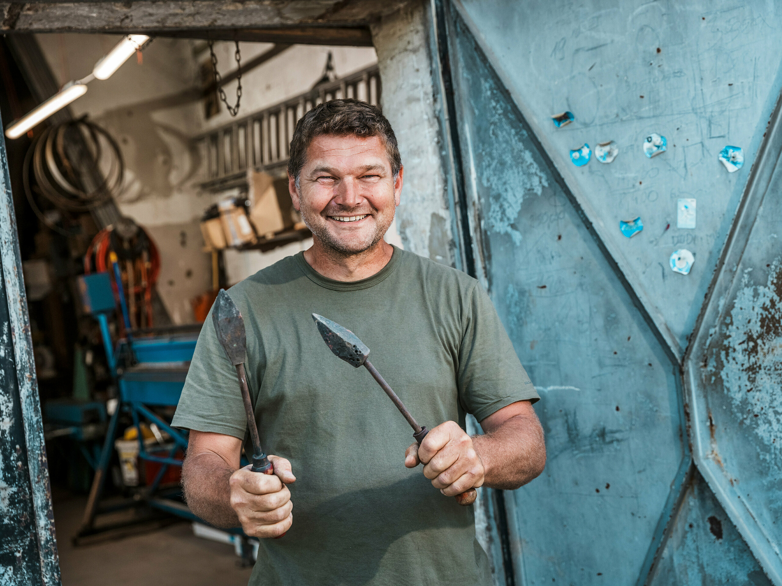 Klampiarsky majster Richárd Tóth vo svojej dielni. Tento zanietený remeselník drží v rukách tradičné náradie a stojí pred otvorenými kovovými dverami. Scéna pôsobí autenticky a zdôrazňuje remeselnú zručnosť a vysokú presnosť – vlastnosti, ktoré boli kľúčové pri spracovaní hliníkových produktov PREFA pre budapeštiansky bytový dom. Surové prostredie dielne podčiarkuje odborné schopnosti, s ktorými boli vyrobené fasádne prvky, atika a obklady balkónov zo systému PREFALZ v odtieni P.10 antracit. Obrázok tak vyzdvihuje spojenie skúseností, materiálovej odbornosti a trvácnych riešení PREFA.