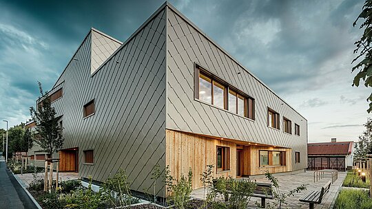 V jemnej večernej atmosfére rozvíja hybridná budova materskej školy v Alzenau svoj plný architektonický účinok. Výrazná fasáda z bronzovo sfarbených PREFA fasádnych šablón 44 × 44 dodáva budove elegantný, odolný vonkajší plášť s jasnou štruktúrou. Na prízemí vertikálne drevené obklady vytvárajú prirodzený, teplý kontrast k hliníku a zvýrazňujú vstupnú časť i chránené pobytové zóny. Zredukované, moderné tvaroslovie harmonizuje s upraveným vonkajším prostredím, kde lavičky, stojany na bicykle a mladé stromy lákajú k pobytu. Obrázok pôsobivo ukazuje, ako trvácne produkty PREFA prispievajú k udržateľnej, funkčnej a zároveň prívetivej vzdelávacej stavbe.