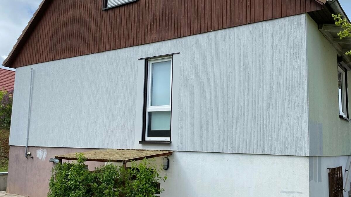 Ein Einfamilienhaus vor der Sanierung mit einer in die Jahre gekommenen Holzfassade im Giebelbereich. Die alte Fassade wurde durch eine hochwertige Aluminiumverkleidung von PREFA ersetzt, die das Gebäude langlebiger und optisch ansprechender macht. Der Vorher-Nachher-Vergleich macht die Unterschiede deutlich.