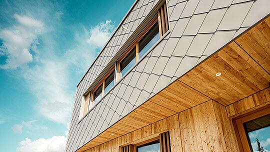 Detailný záber detského zariadenia v Alzenau ukazuje precízne vyhotovenie a vysokokvalitnú kombináciu materiálov z bronzovo sfarbených PREFA fasádnych šablón 44 × 44 a prírodného, neupravovaného dreva. Diagonálna štruktúra hliníkovej fasády vytvára výrazný šupinový vzor, ktorý sa harmonicky prepája s vertikálnymi drevenými lamelami pri oknách a jemne spracovaným dreveným podhľadom. Záber z nadhľadu od zeme (tzv. žabia perspektíva) zdôrazňuje presné vedenie línií a remeselnú kvalitu prevetrávanej zavesenej fasády. Tento detailný pohľad podčiarkuje dizajnovú silu a dlhú životnosť produktov PREFA – ideálne pre udržateľné architektonické projekty, ktoré spájajú estetiku, funkčnosť a ekológiu na najvyššej úrovni.