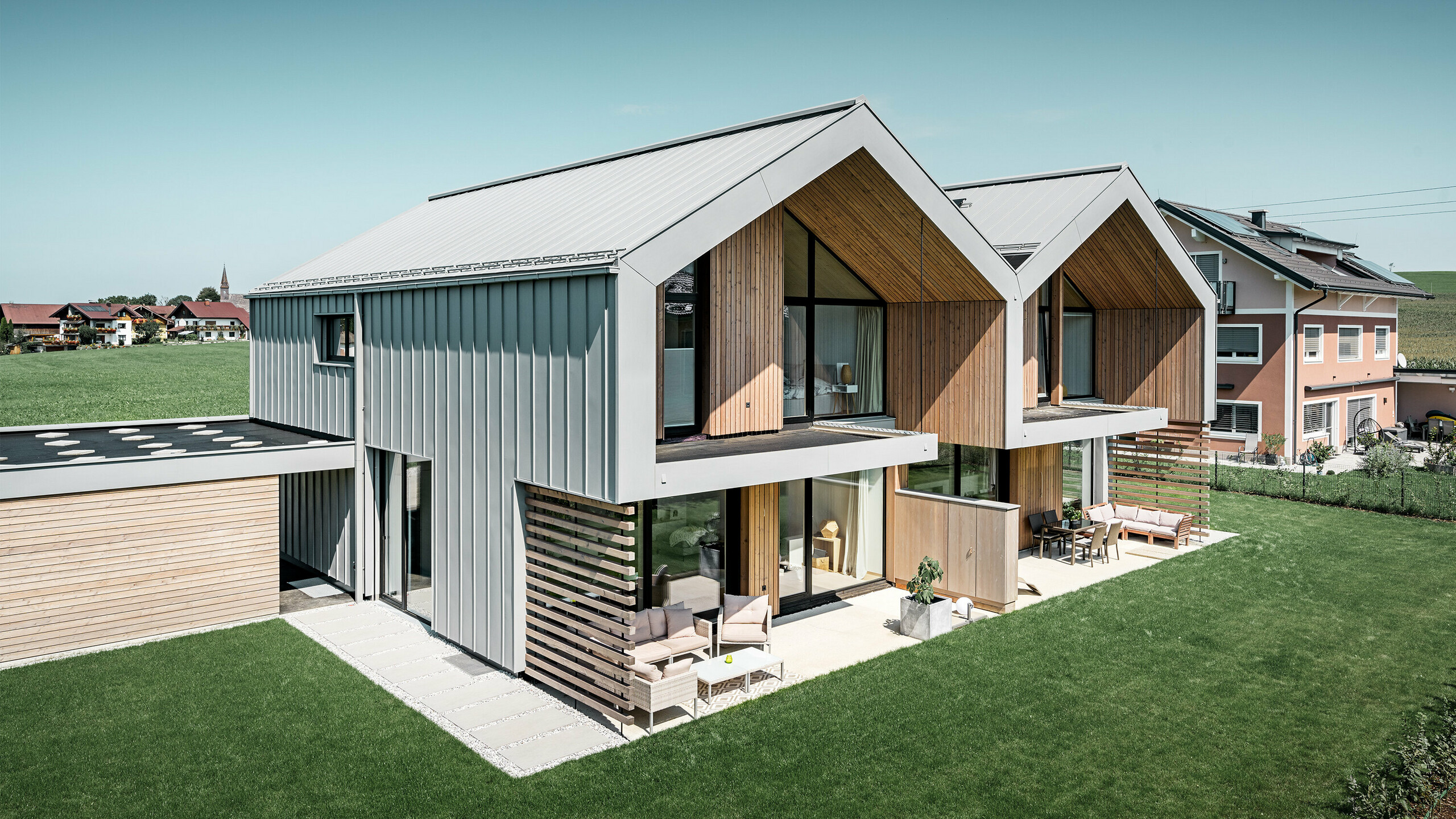 Moderný dvojdom s hliníkovým plášťom. Plechová strecha a plechová fasáda sú vyrobené z PREFALZ v odtieni patina šedá. Ľahký obklad stojatej drážky v kombinácii s drevenými prvkami harmonicky zapadá do okolia. Odvodnenie strechy prebieha cez hranatú rúru PREFA a je tiež vo farbe patina šedá.